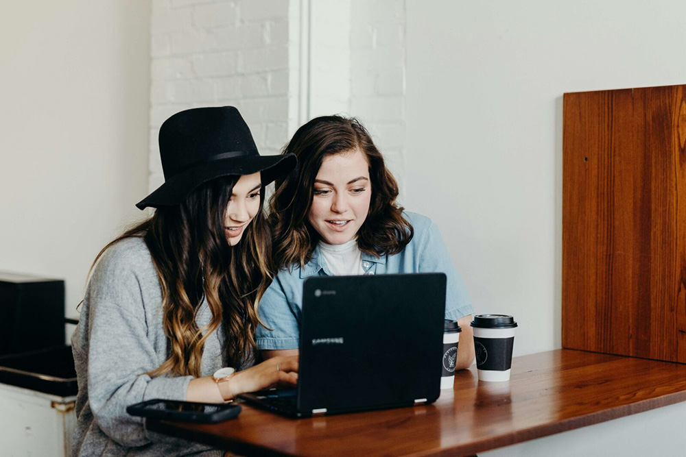 Two woman sitting at a coffee table with coffee cups while looking at a computer screen. If you suffer from work anxiety, work with an anxiety therapist in NYC, NY today! Call now. 