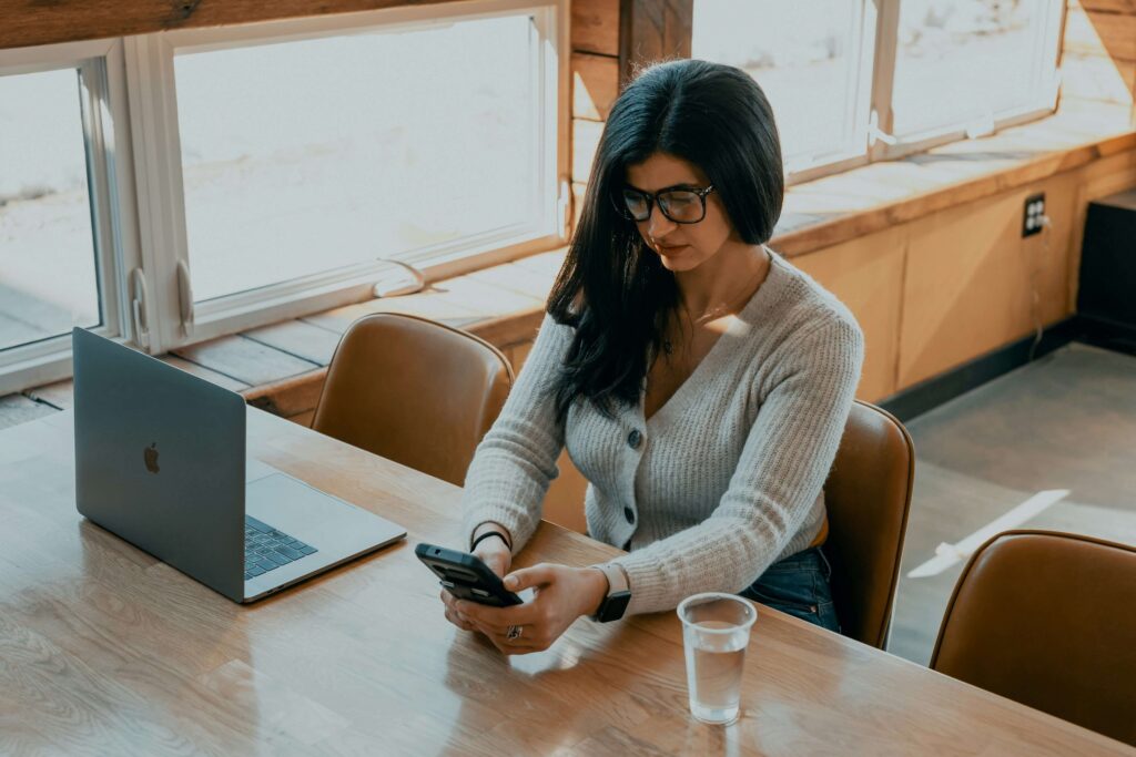 A woman with brown hair typing on her phone & sitting at a large table with a laptop. If you want to learn about the benefits of anxiety therapy in NYC, NY then contact us today! We can help you manage your anxiety.