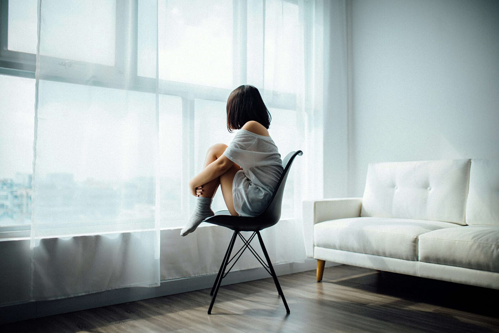 A woman sitting alone in a chain in front of a large window looking out. Representing how anxious attachment can be lonely. Get started with an anxiety therapist in NYC, NY to help!