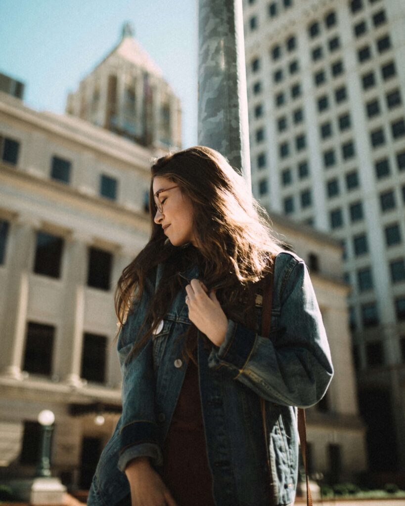 A woman standing in a city smiling to the side & closing her eyes. Representing how secure attachment looks like maintaining a positive self-image. Learn more in anxiety counseling NYC. 