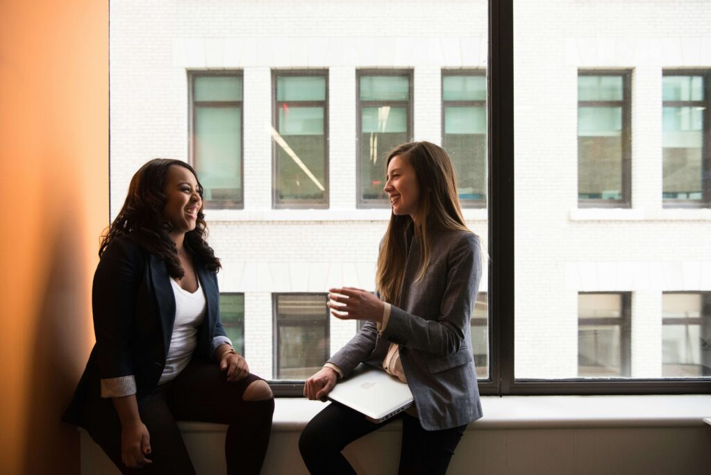 Two NYC professionals talking near a large window while smiling. With the help of an anxiety therapist in NYC, you can beat corporate burnout. Get started today.