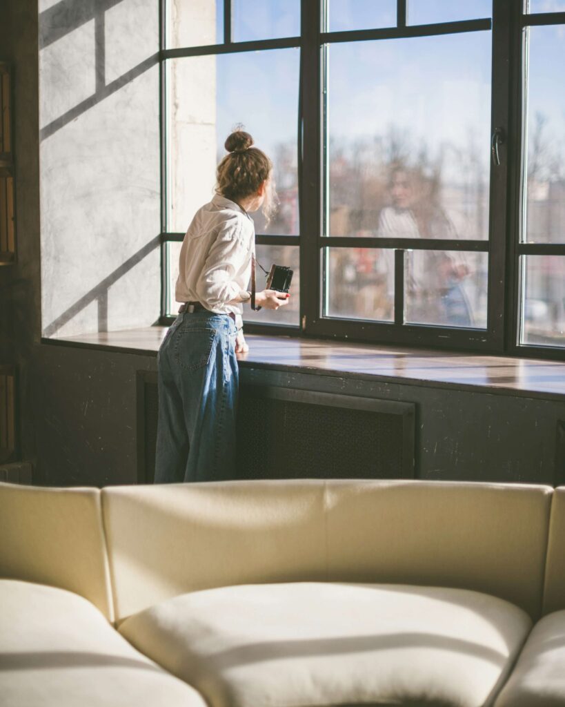 A woman standing near a large window holding a coffee mug. Did you know anxiety counseling in NYC, NY can help alleviate burnout symptoms? Get started with anxiety therapy today!
