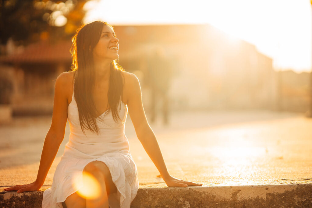 A young woman wearing a white dress smiling & looking away while sitting on a ledge. Representing how working with an anxiety therapist in NYC, NY can help you with procrastination. Start your journey today!