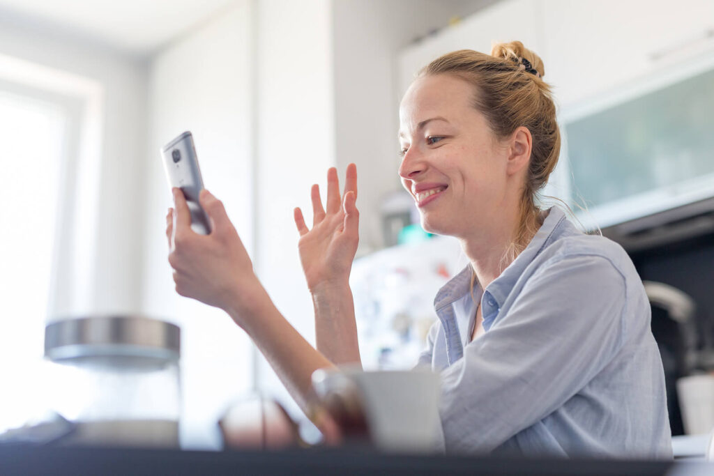 A woman holding a phone while waving & smiling at the online therapist. Representing how online anxiety therapy in NYC, NY is great for busy women. Fit an anxiety therapy session into your own schedule at any time!